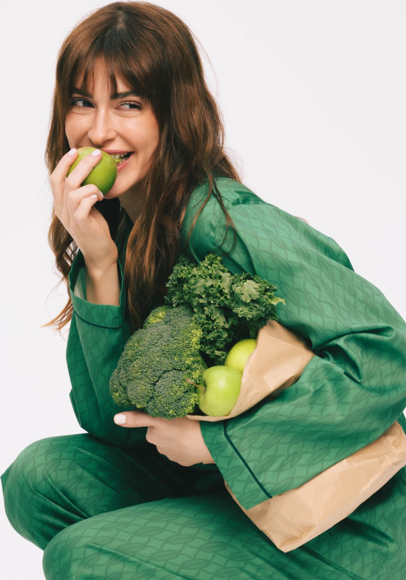 Woman in green biting an apple, holding a bag of vegetables